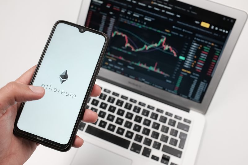 A person holds a cell phone with an Ethereum logo, while a laptop displaying stock charts appears in the background.