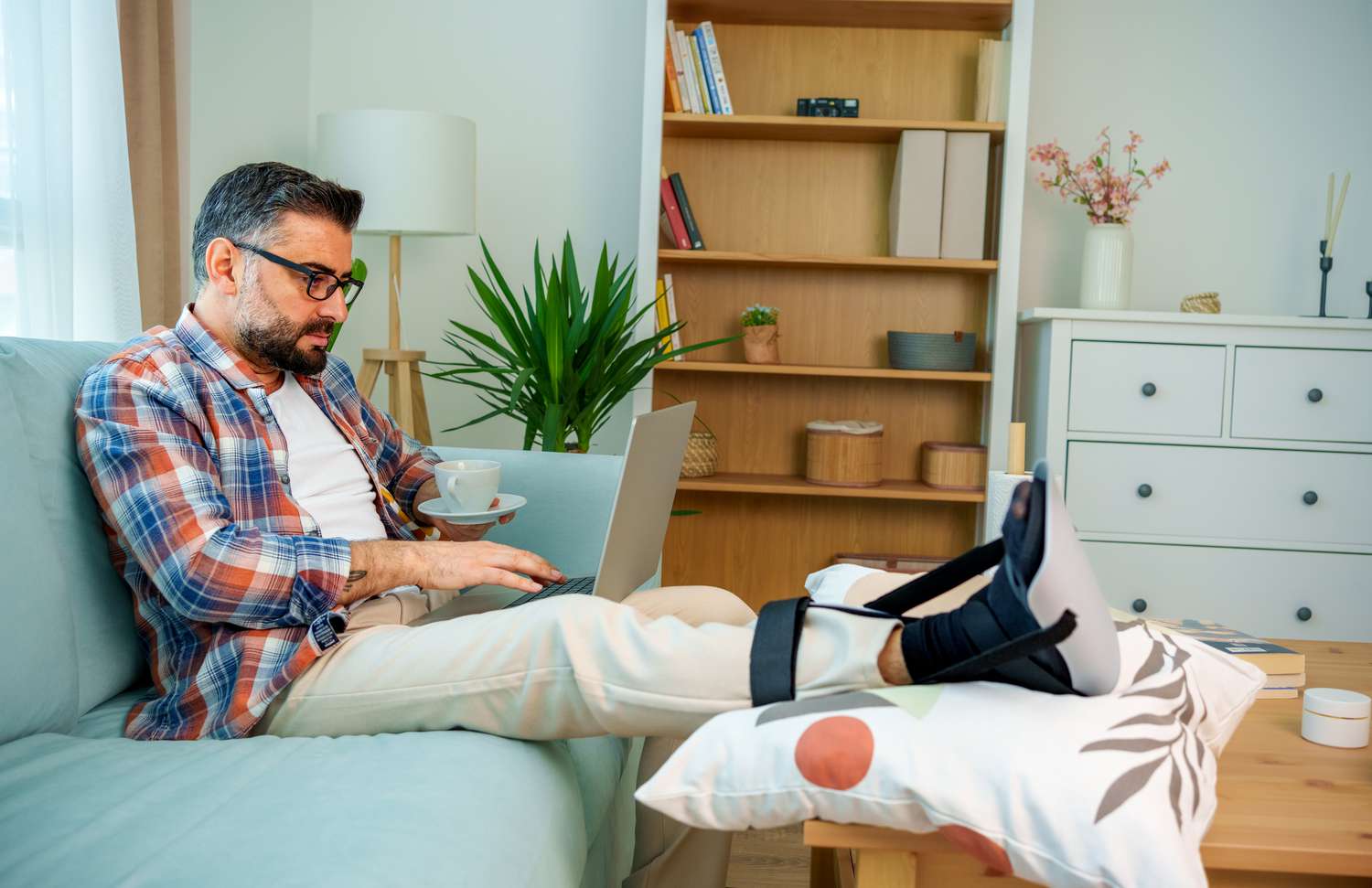 man working on laptop in home stretches broken foot in a cast on a pillow placed on the coffee table