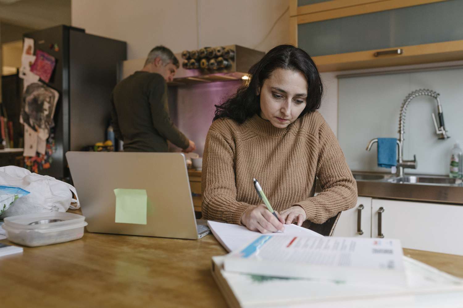 Mature woman working on the paper for her loan in the kitchen, husband cooking behind
