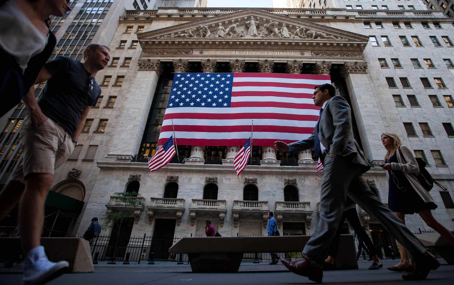 Pedestrians walk past a large American flag hanging outside the New York Stock Exchange building.
