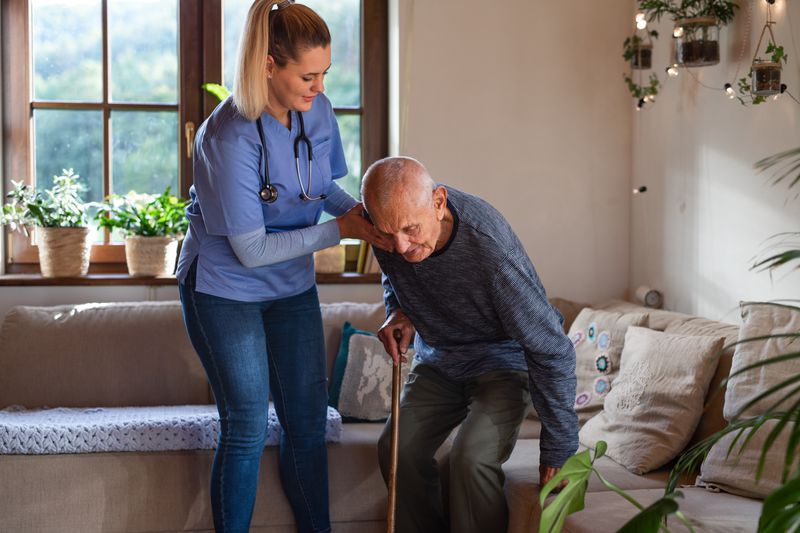 Nurse helping senior patient to stand up from the sofa in a long-term care facility included as an insurance rider.