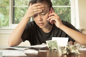 A stressed woman looks at bills on a table, holding a cell phone to her ear.