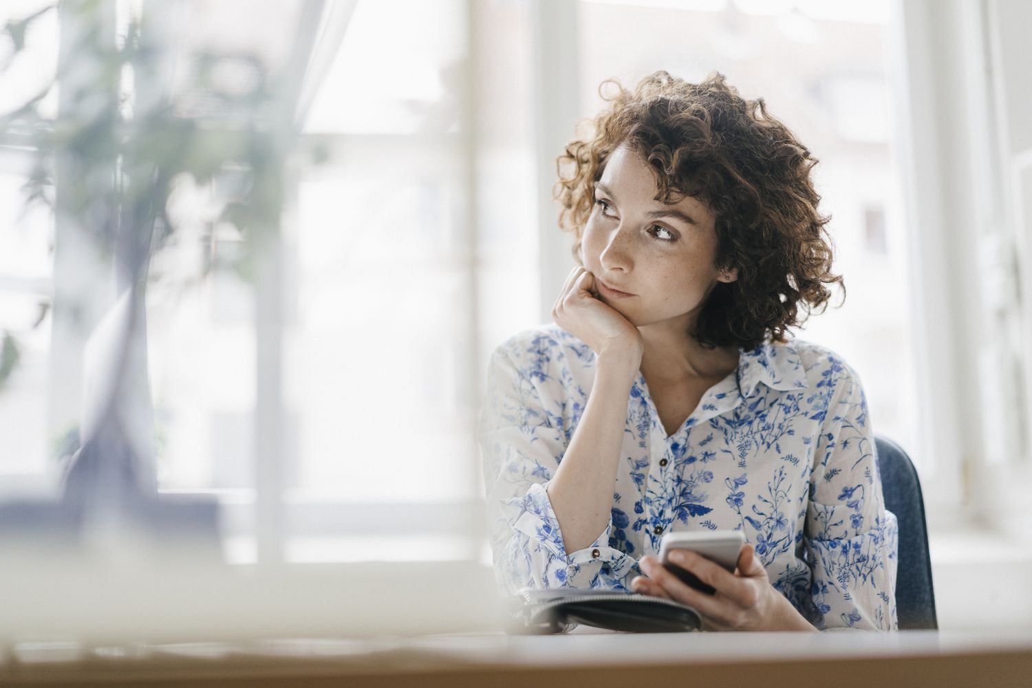 A woman looking contemplative while holding up a smart phone and sitting in a living room