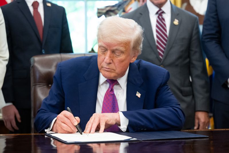 Donald Trump sitting at a desk, signing a document, with others standing in the background