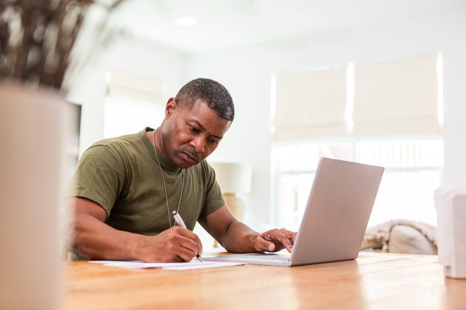 A man sits at a table in his home with a laptop and a pen and paper 