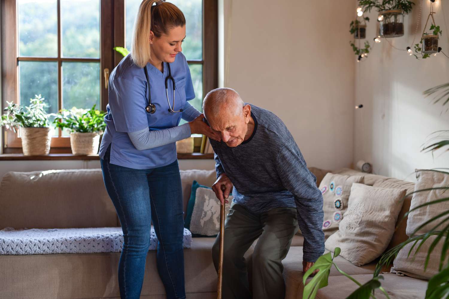 Nurse helping senior patient to stand up from the sofa in a long-term care facility included as an insurance rider.