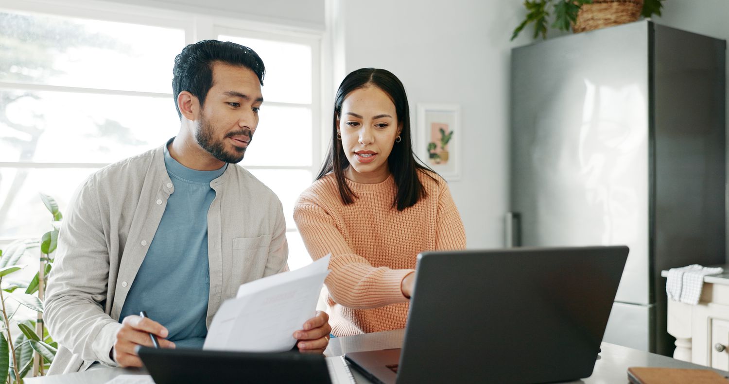 Young couple sitting in their kitchen and looking at mortgage documents and rates on a laptop