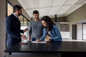A man and a woman smile while signing papers on a kitchen counter with a real estate agent. 