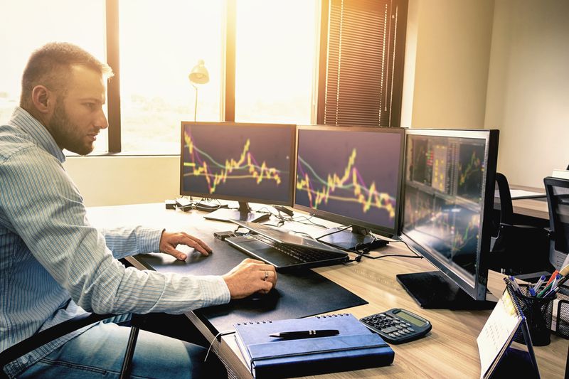 Young trader is working at stock market with three computer screen on his office desk.