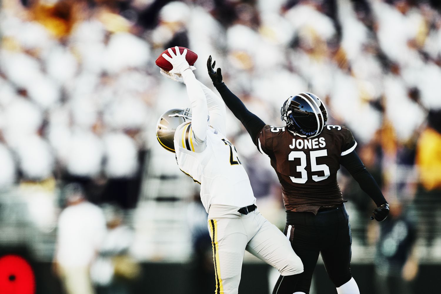 Professional football players on the field with one catching a ball and the other reaching for it.
