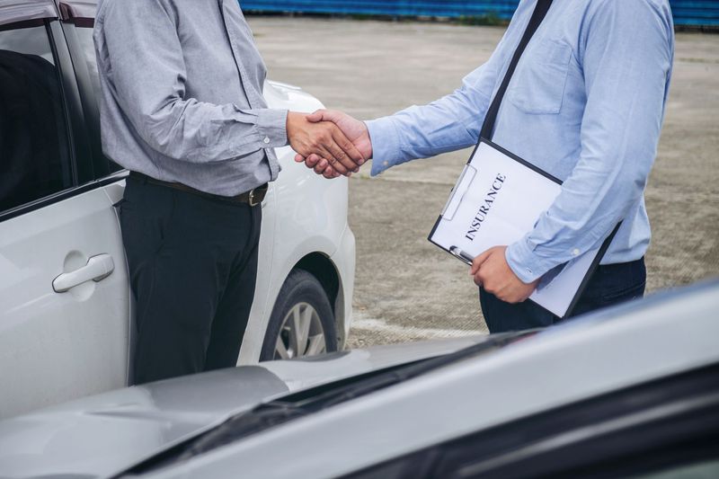 Two men shaking hands near two cars, one holds document labeled "Insurance"