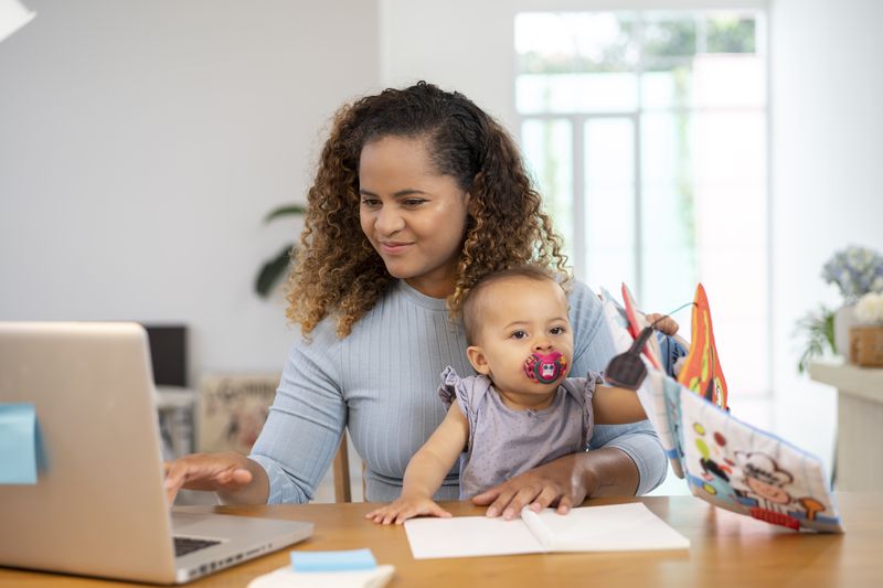 Young mom sitting at dining room table with baby on her lap, checking something on her laptop