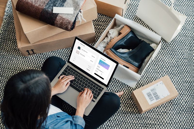 High-angle view of young woman on computer surrounded by packages, one of them open with a pair of boots inside