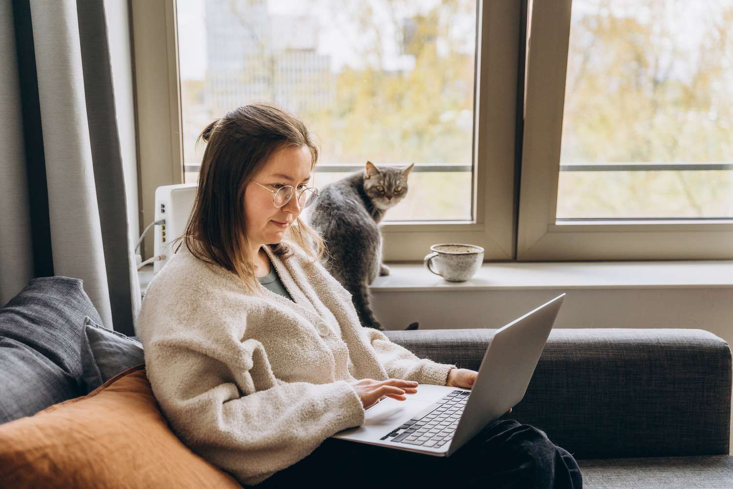Young woman working at home remotely using a laptop while sitting on the sofa. Her domestic cat is sitting next to her