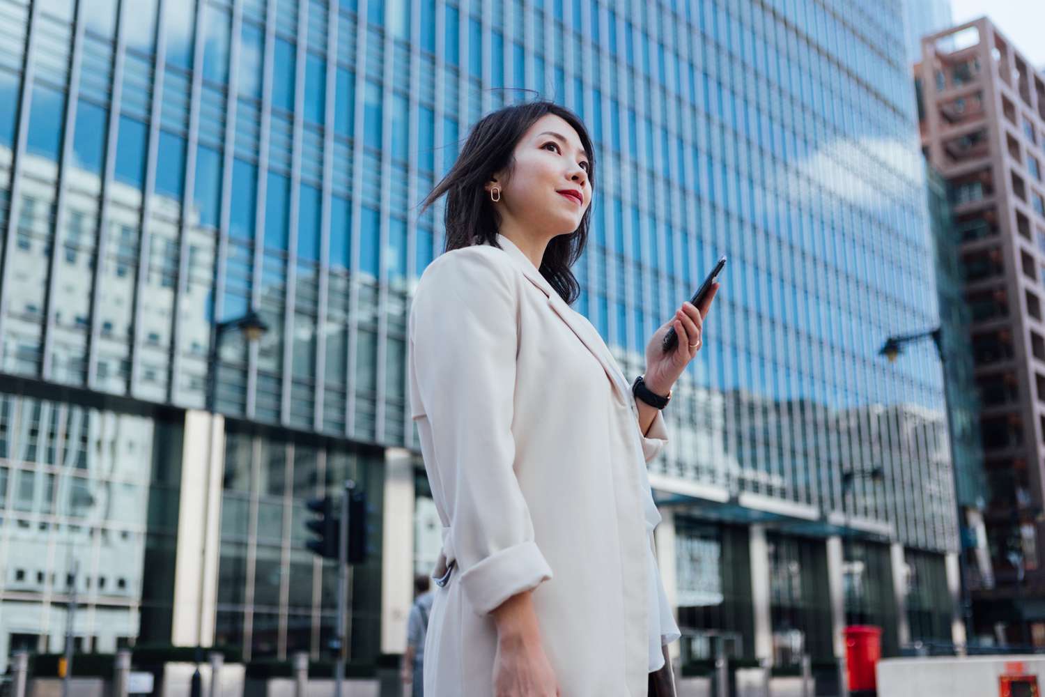 Low-angle view of an Asian businesswoman looking away, holding a smartphone, standing against modern buildings in the city. 