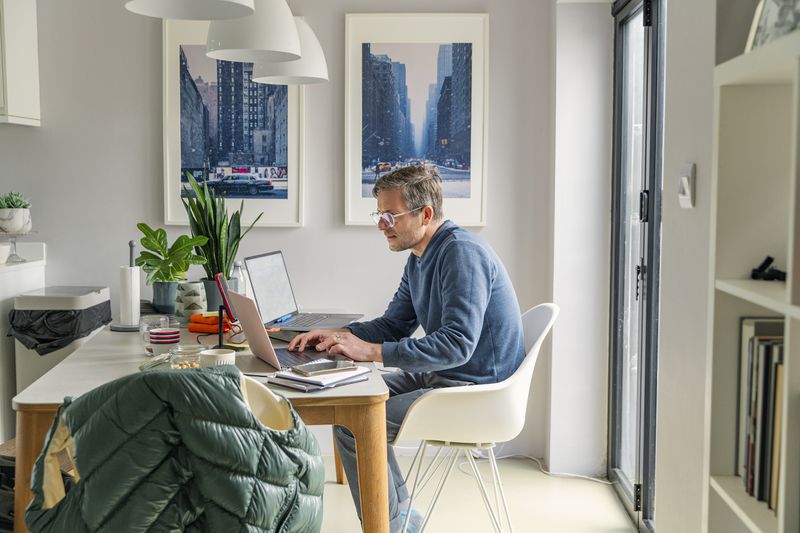 White man with glasses sitting at a desk in office-like at home setting with two laptops. 
