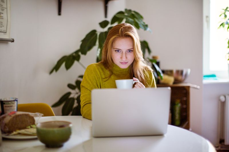 Young woman sitting at table looking at her laptop and drinking coffee.
