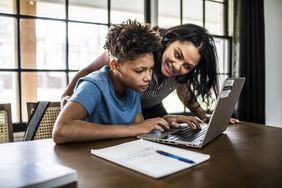Adult woman helping younger man on laptop at a dining room table.