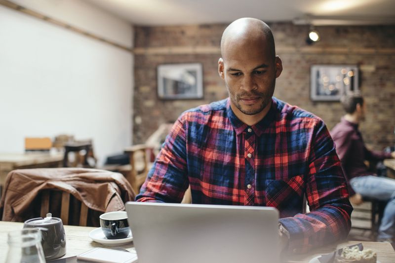 A man in a plaid shirt looks at his laptop in a coffee shop