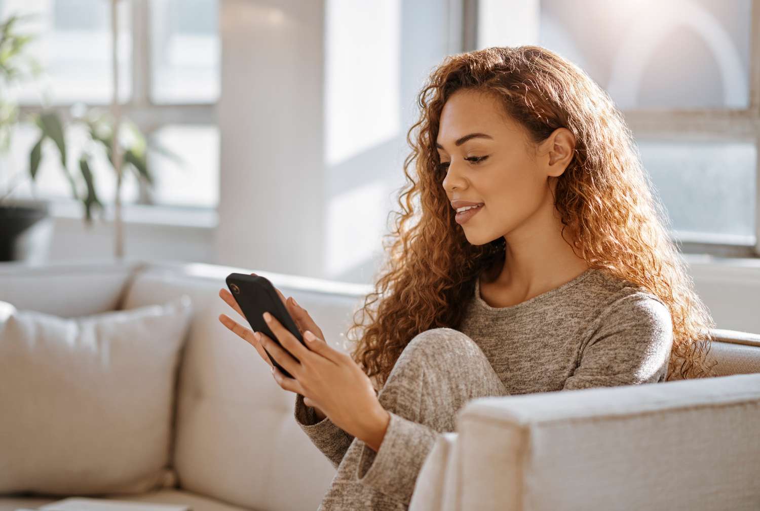Young woman sitting on her living room couch and looking with a smile at something on her smartphone