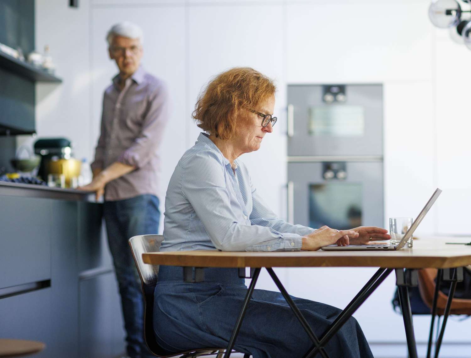 Woman working on a laptop while a man is looking at her from the kitchen