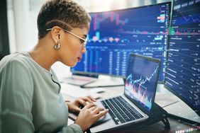 Female traders sitting at desk working on a laptop. Several monitors showing financial data are behind the laptop.