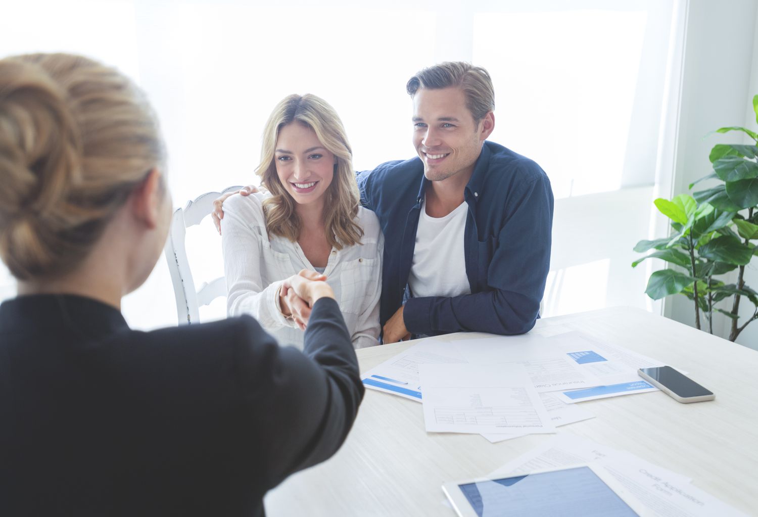 Real estate agent with couple shaking hands closing a deal. There is a digital tablet on the table. Couple are casually dressed. They are looking happy and smiling. Over the shoulder view inside a home.