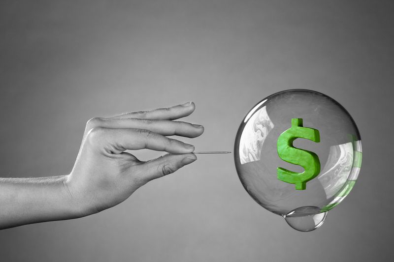 A hand about to burst a money bubble with a pin.