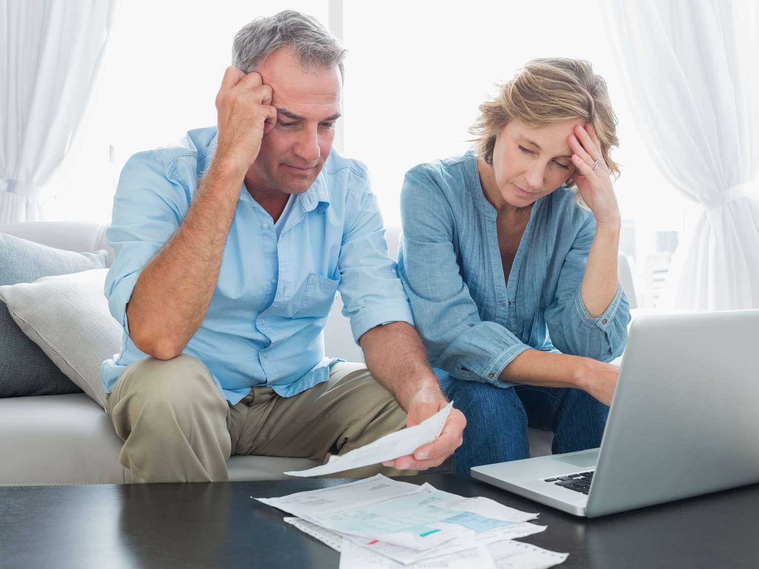 Couple in their 50s sitting on their sofa and looking with concern at mortgage documents and a laptop