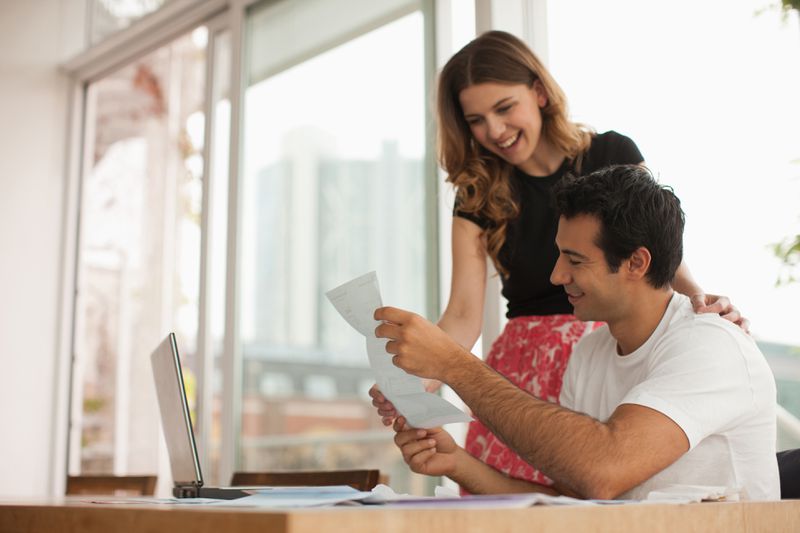 Two people in a kitchen in front of a laptop holding a piece of paper