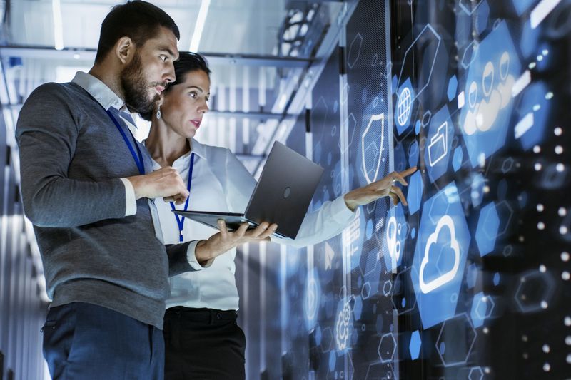 Male IT specialist holds laptop and discusses work with female server technician.