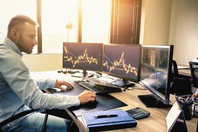 Young trader is working at stock market with three computer screen on his office desk.