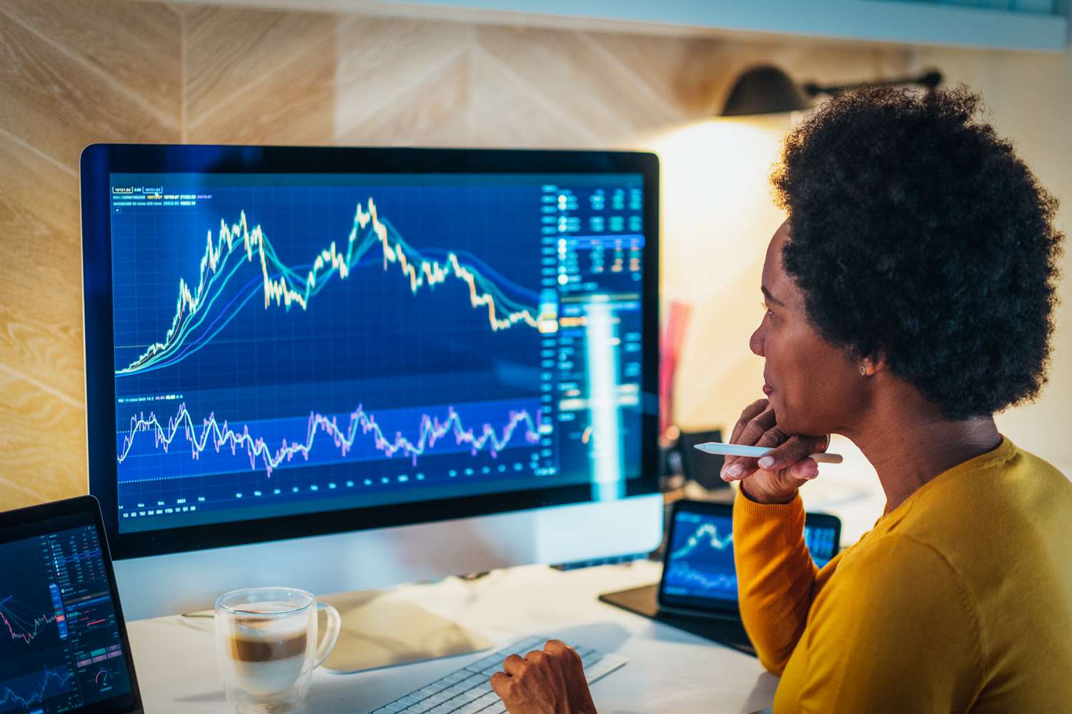 A woman sits at a desk with a cup of coffee in front of a large computer screen displaying financial stock charts. 
