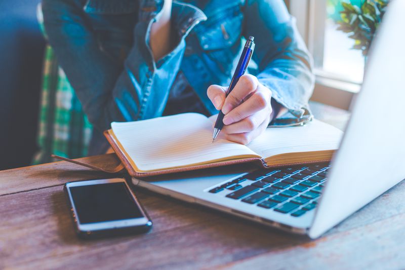 Student at desk, writing in journal on top of open laptop