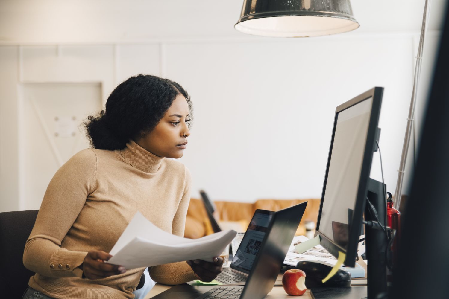 Focused female IT professional holding document while looking at computer in creative office