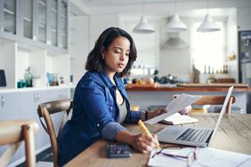A woman reviews paperwork and uses a laptop in her kitchen
