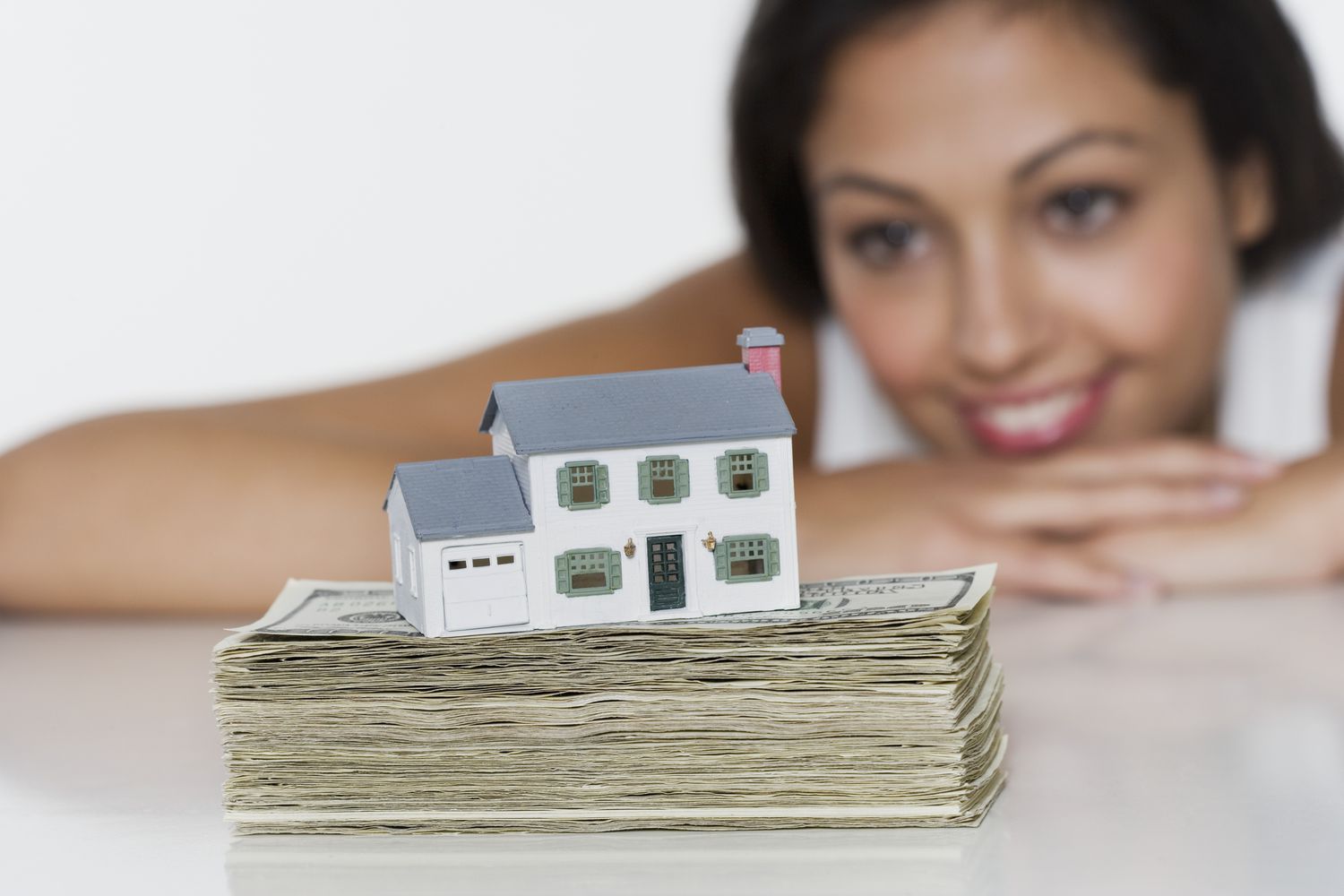 A woman looks at a pile of money with a miniature house on top.