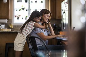 Daughter hugging mom from behind while the other one is on the phone looking for ways to save their house from foreclosure.