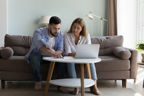 Millennial couple sitting on living room sofa and studying laptop and paperwork together