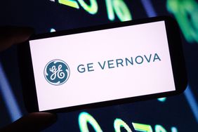 The blue-green GE Vernova is seen on a phone screen, in front of a screen with green numbers in the background.
