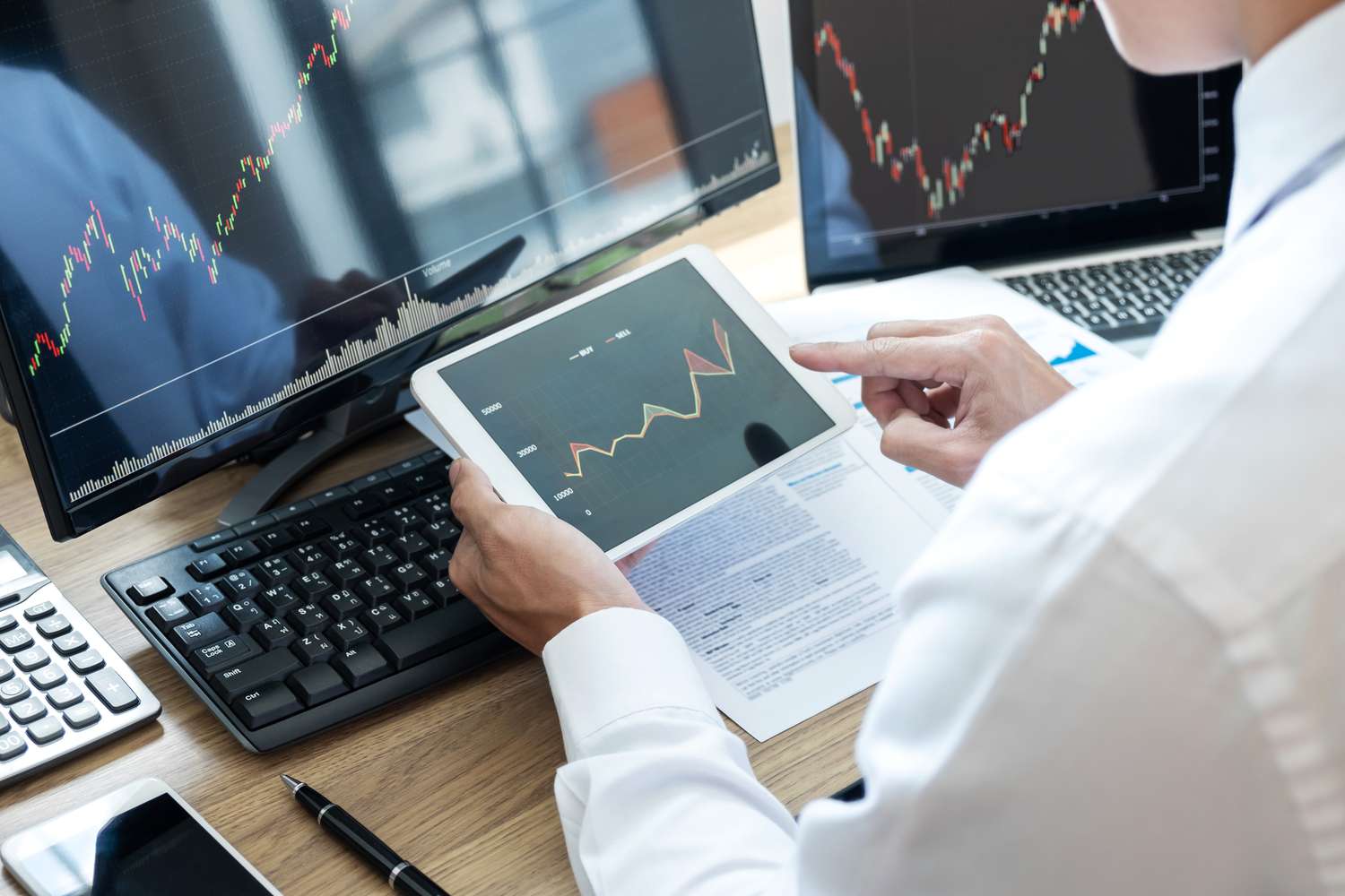 A close-up of a man holding a tablet with a stock chart, while sitting in front of two computer screens.