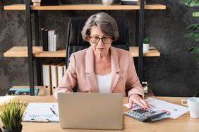 An older woman working at a desk with a laptop and calculator