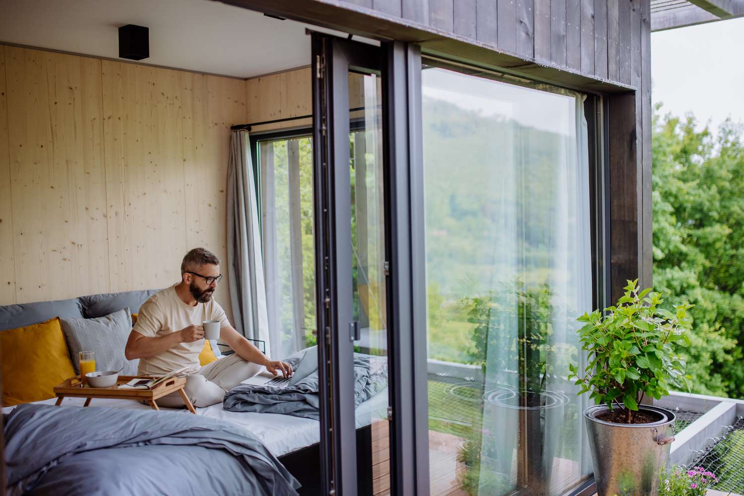 A man sitting on a bed near a large sliding glass door, in a modern and well-lit room