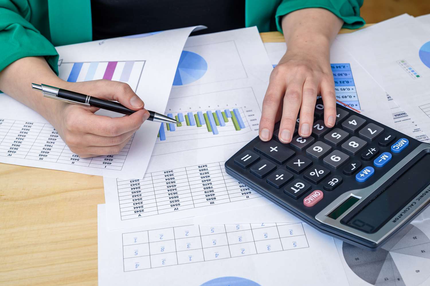 Desk strewn with documents and a pair of hands holding a pen and touching a calculator.