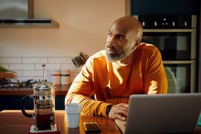 A person sitting at a kitchen table with a laptop and a French press coffee maker
