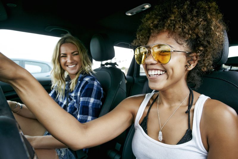Two happy women driving in a car, smiling. Driver has curly brown hair and sunglasses.