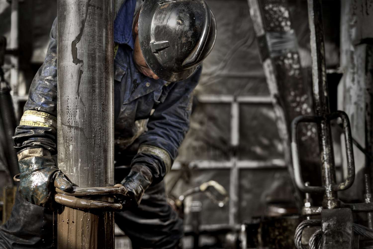 A worker wears a hardhat and heavy gloves as they tighten a cable around the drill head of an oil rig.