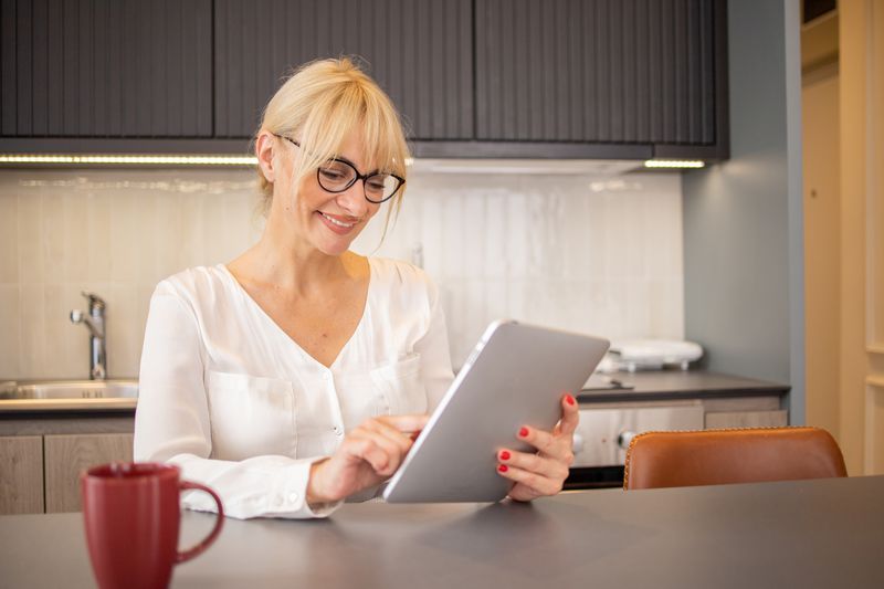 Woman in her 50s looking happily at a tablet while sitting at her kitchen counter