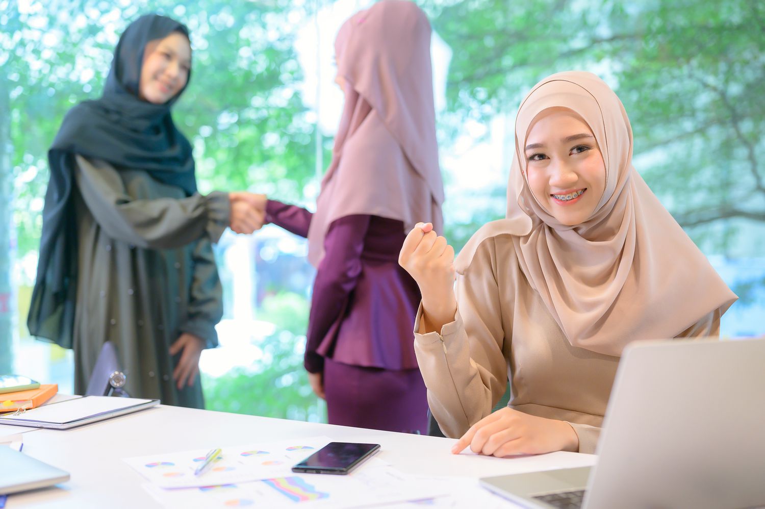 Woman sitting at a desk, looking up and smiling. Two women shake hands behind her.