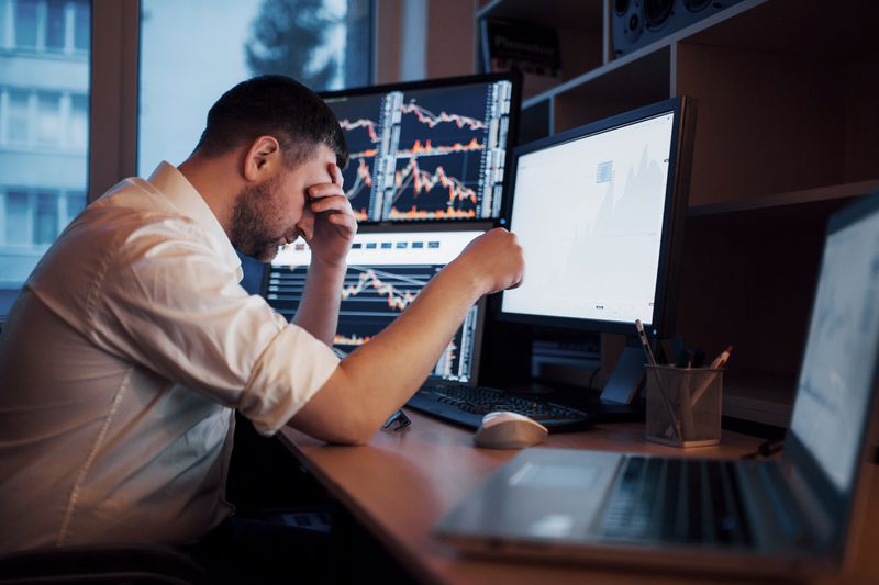 Stressed stock trader looking frustrated in front of trading screens after a day of yo-yo market that leads to losses.